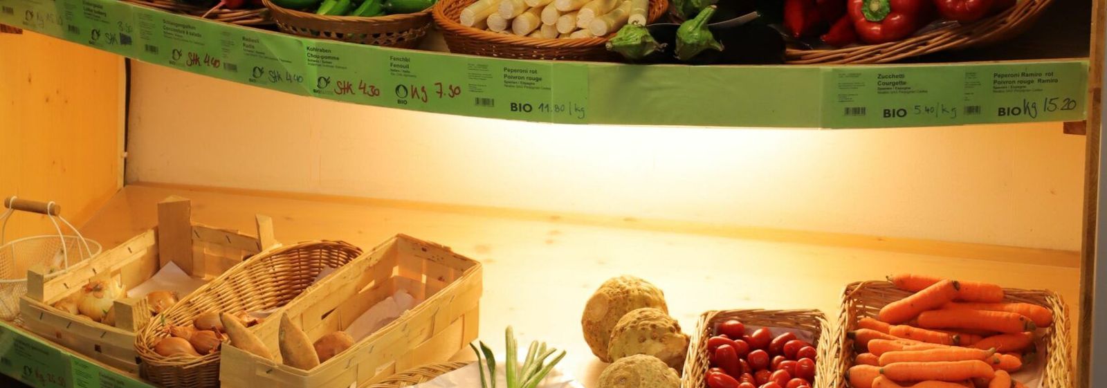 Regalbretter im Laden mit Weidenkörben voller Obst und Gemüse, darüber Reihen beschrifteter Vorratsgläser